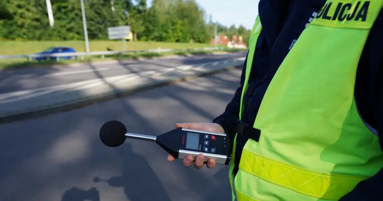 Policjant w kamizelce odblaskowej trzyma miernik dźwięku, by sprawdzić, jak zmierzyć głośność wydechu pojazdu.