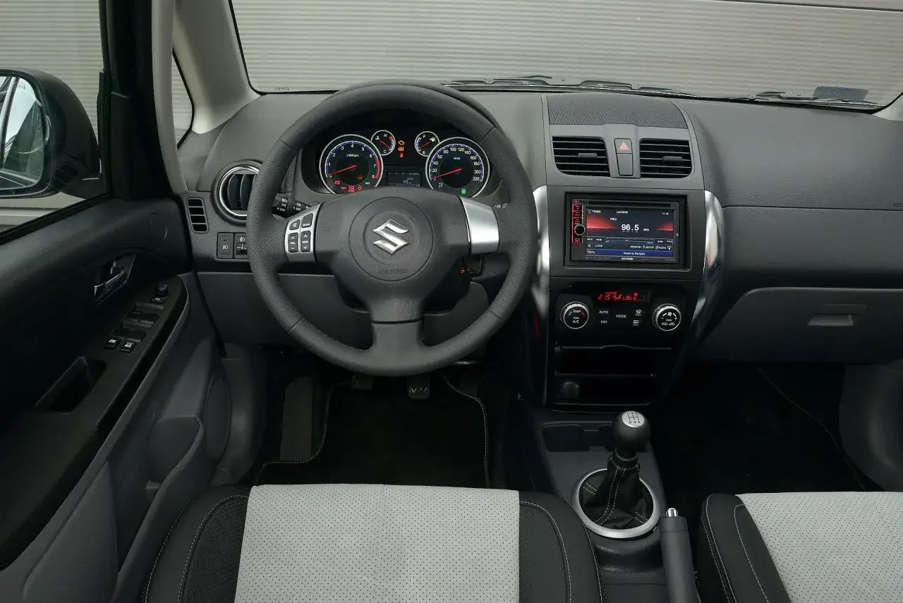 Wnętrze Suzuki SX4 1.6 benzyna: kierownica, deska rozdzielcza z radiem i klimatyzacją, dźwignia zmiany biegów.
